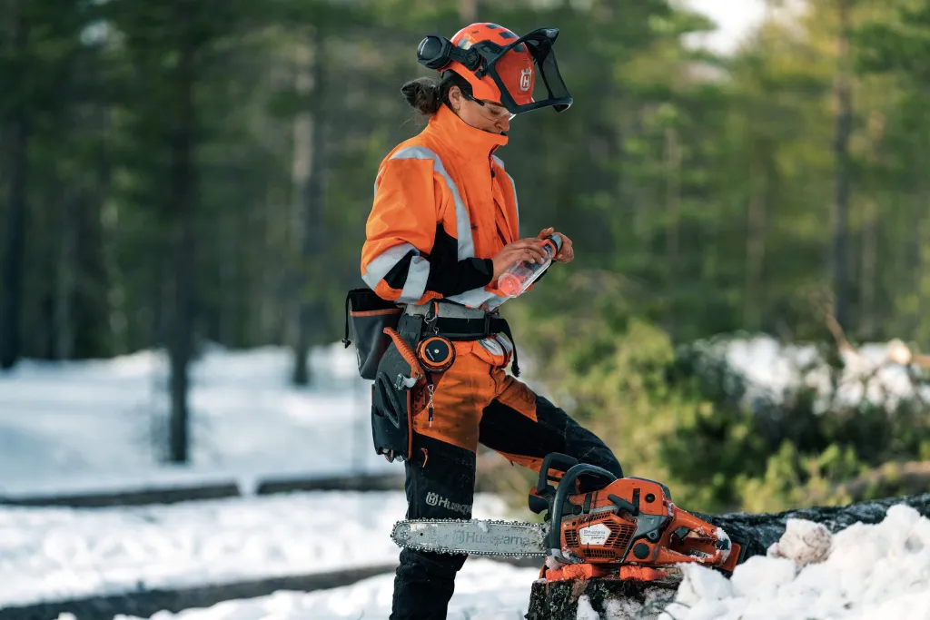 Motorsågsförare i skyddskläder och hjälm med visir förbereder motorsåg på stubbe i vintrig skog
