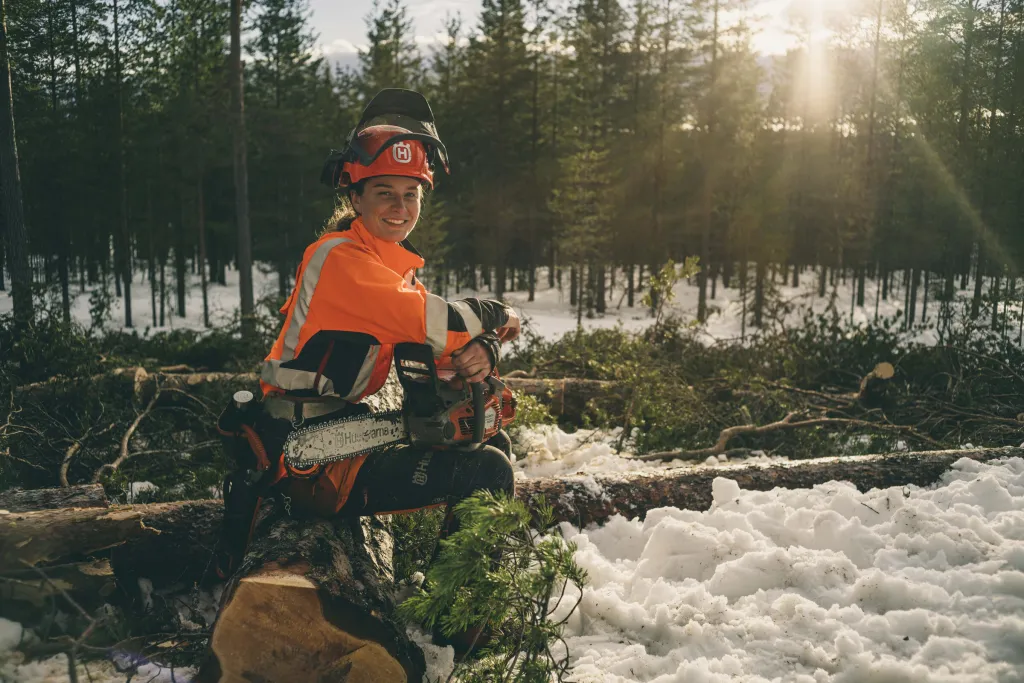 Leende motorsågsförare i skyddsutrustning sitter på en stock med motorsåg i snöig skog