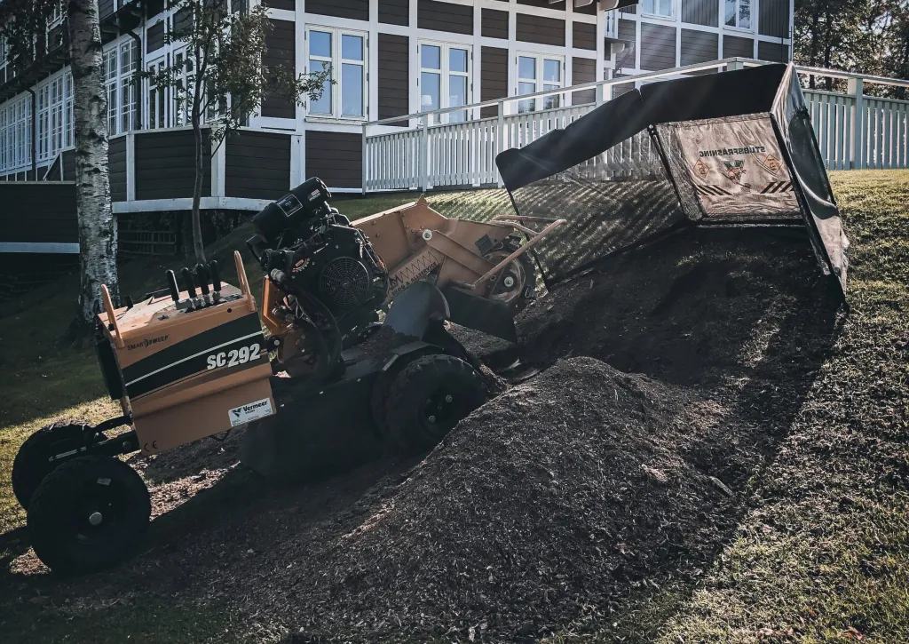 Stubbfräs i arbete på grässlänt framför stort trähus, stubbfräsning i Karlsborg